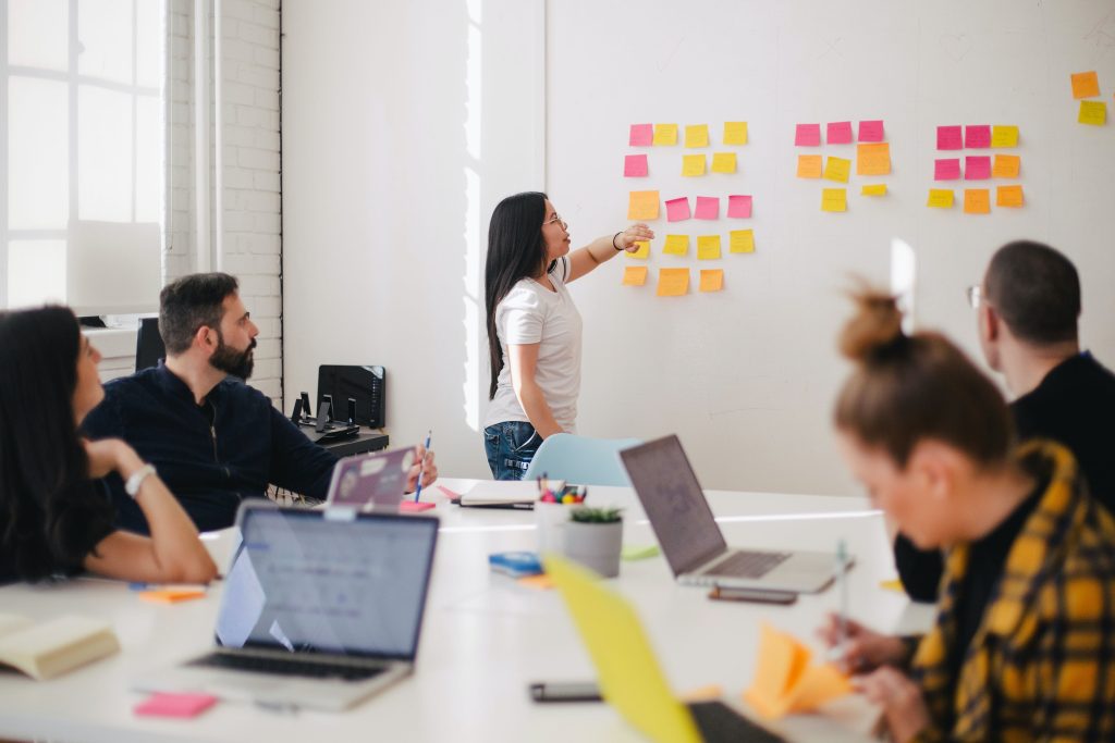 Team meeting, people sat around table looking at woman sticking notes to wall