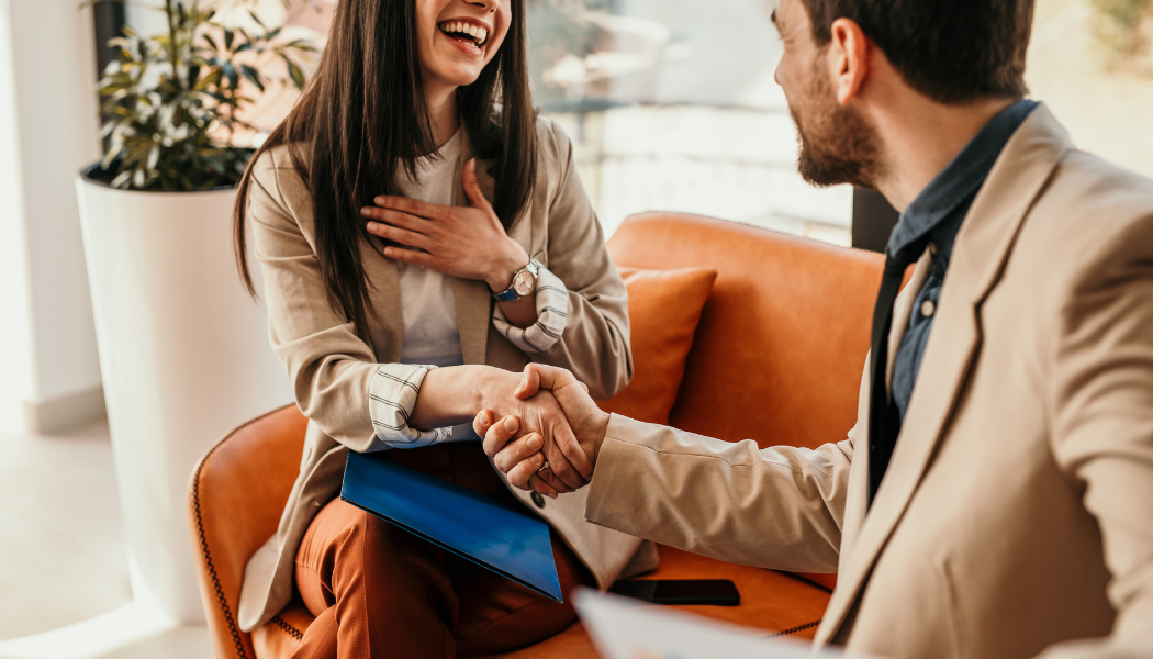 Two people in business attire sit on a sofa, smiling and shaking hands, with a plant and window in the background.