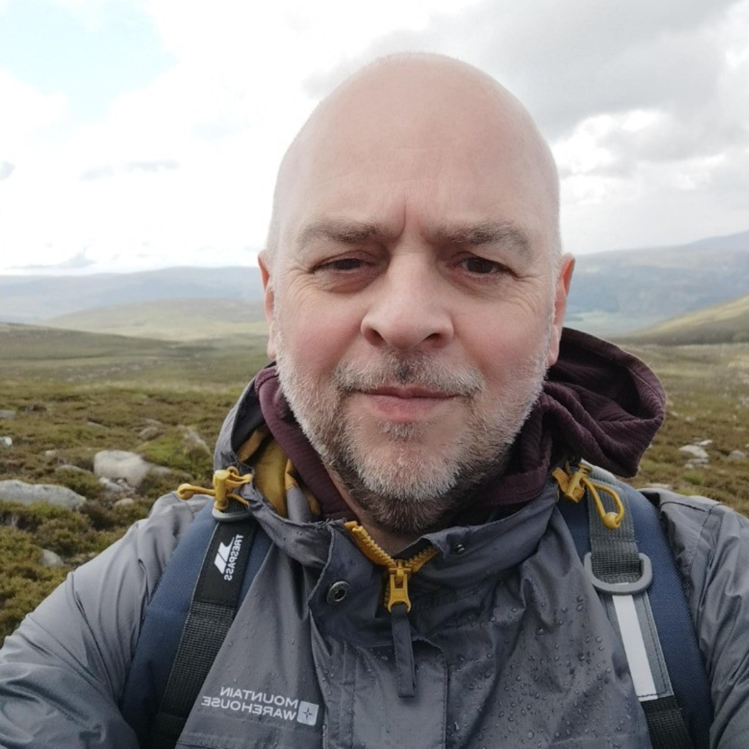 jason matthews A man wearing a grey jacket and rucksack takes a selfie outdoors in a hilly, grassy landscape under a cloudy sky.