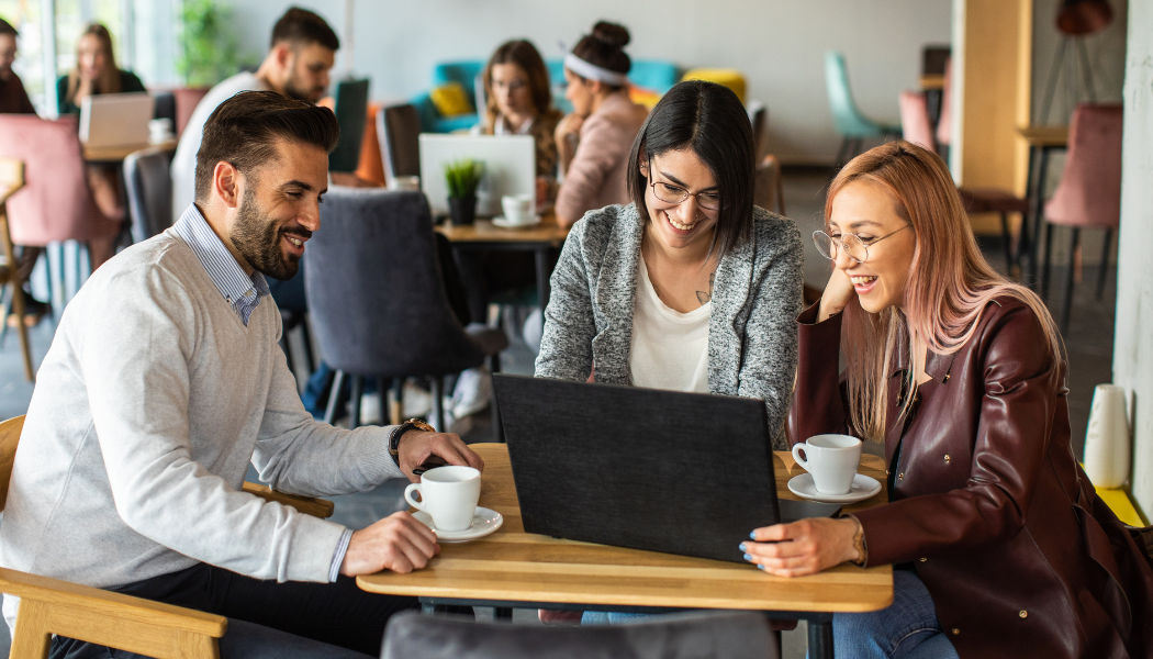 Three people sit at a café table, looking at a laptop and smiling, with coffee cups in front of them; other people work in the background.