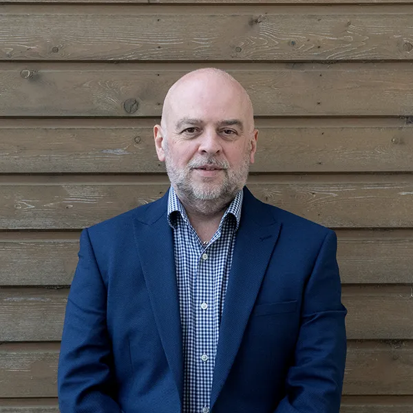 Jason Matthews A middle-aged man with a bald head and beard, wearing a navy blue suit and checked shirt, stands in front of a wooden plank wall.