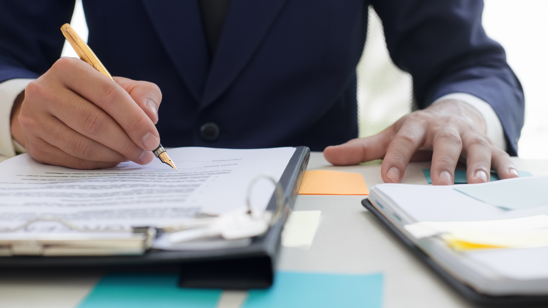 A person in a suit writes on a document at a desk, with a folder, notebook, and scattered sticky notes visible.