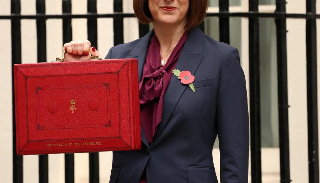 A person in a navy suit holds a large red briefcase labelled "Chancellor of the Exchequer" outside, standing in front of black railings.