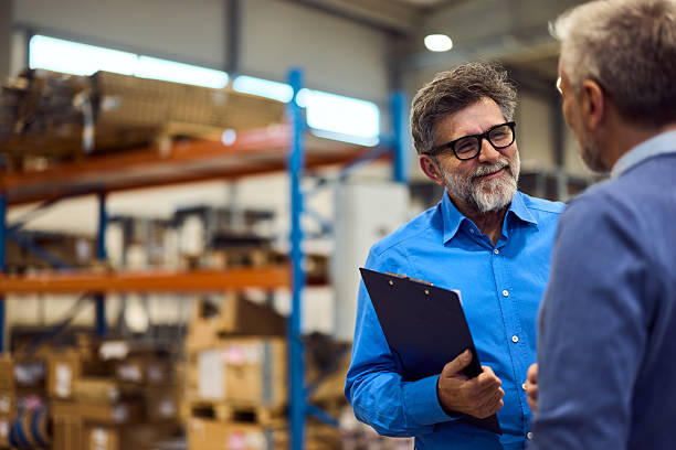 A man in a blue shirt holding a clipboard discusses how to create a job description with another man in a warehouse filled with shelves and boxes.