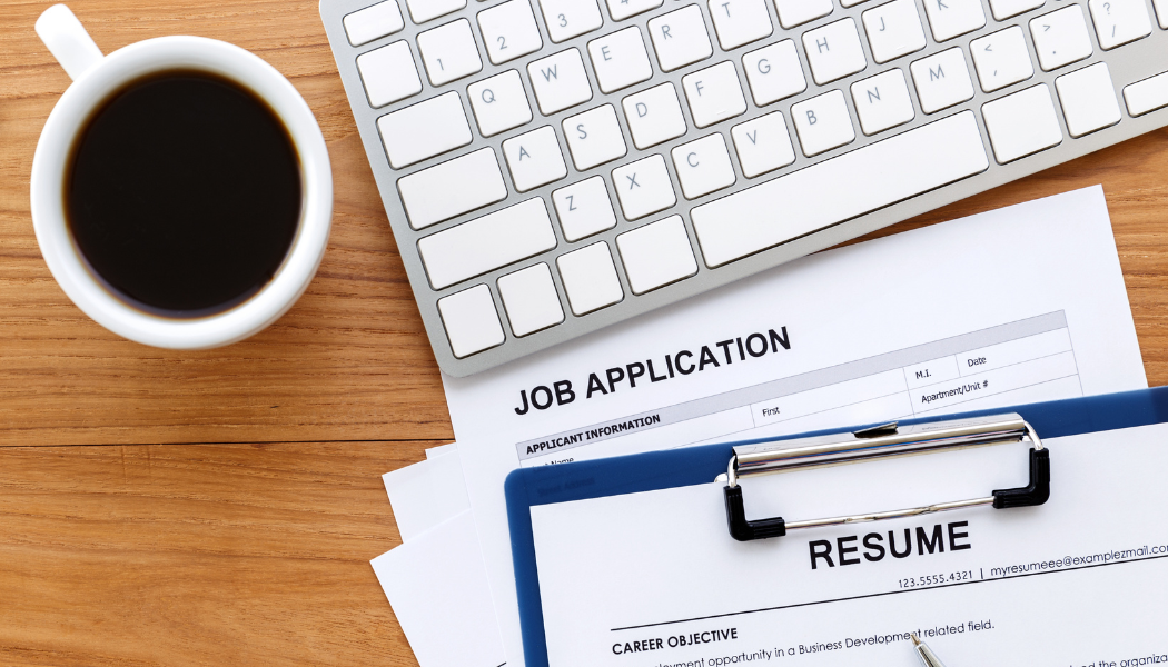 A cup of coffee, a computer keyboard, and documents labelled “Job Application” and “CV” on a wooden desk.