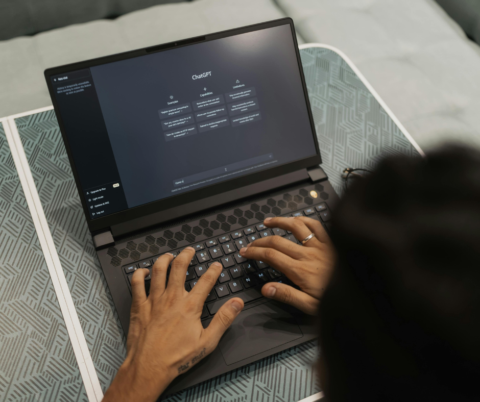 A person types on a laptop displaying the ChatGPT interface while seated at a patterned table, searching for how to create a job description.
